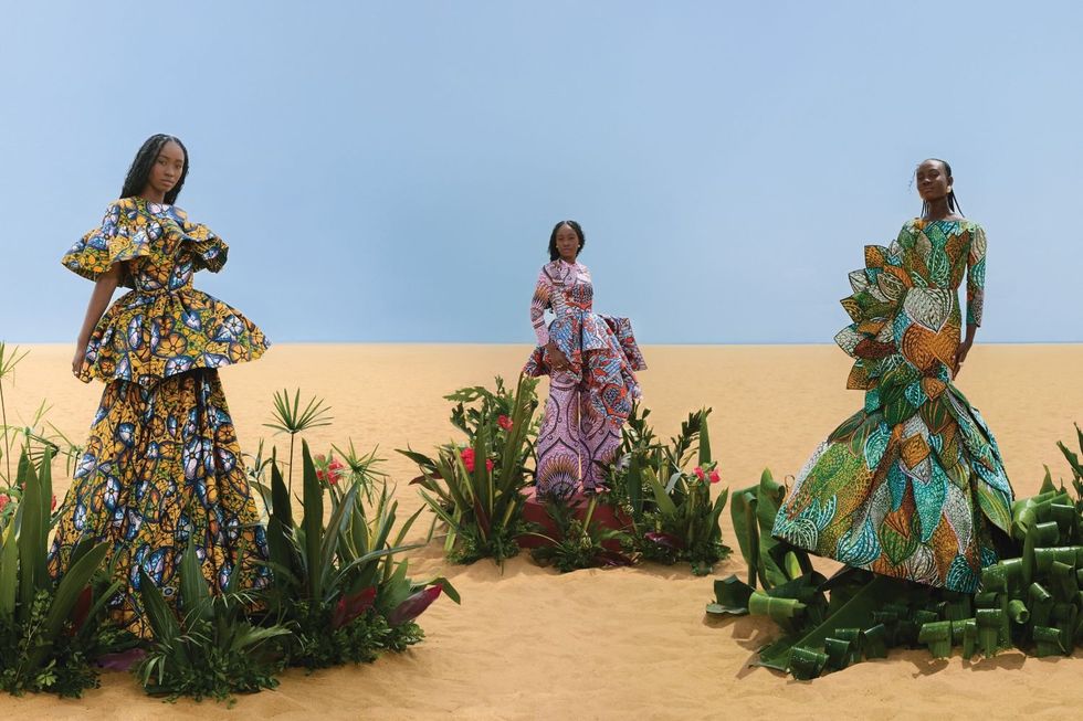 Three models in wax print dresses stand in the middle of colorful shrubs.