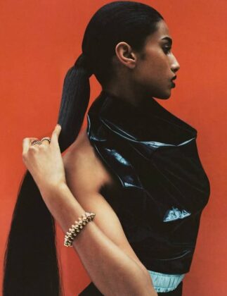 Imaan Hammam wearing a dark practical jacket, standing with her back to the camera, looking out over a still glacial lake reflecting a snow-capped mountain range at dawn.