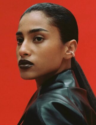 Imaan Hammam wearing a dark practical jacket, standing with her back to the camera, looking out over a still glacial lake reflecting a snow-capped mountain range at dawn.