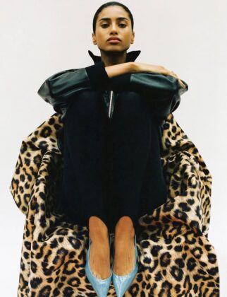 Imaan Hammam wearing a dark practical jacket, standing with her back to the camera, looking out over a still glacial lake reflecting a snow-capped mountain range at dawn.