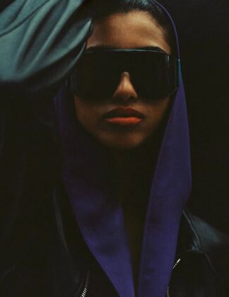 Imaan Hammam wearing a dark practical jacket, standing with her back to the camera, looking out over a still glacial lake reflecting a snow-capped mountain range at dawn.